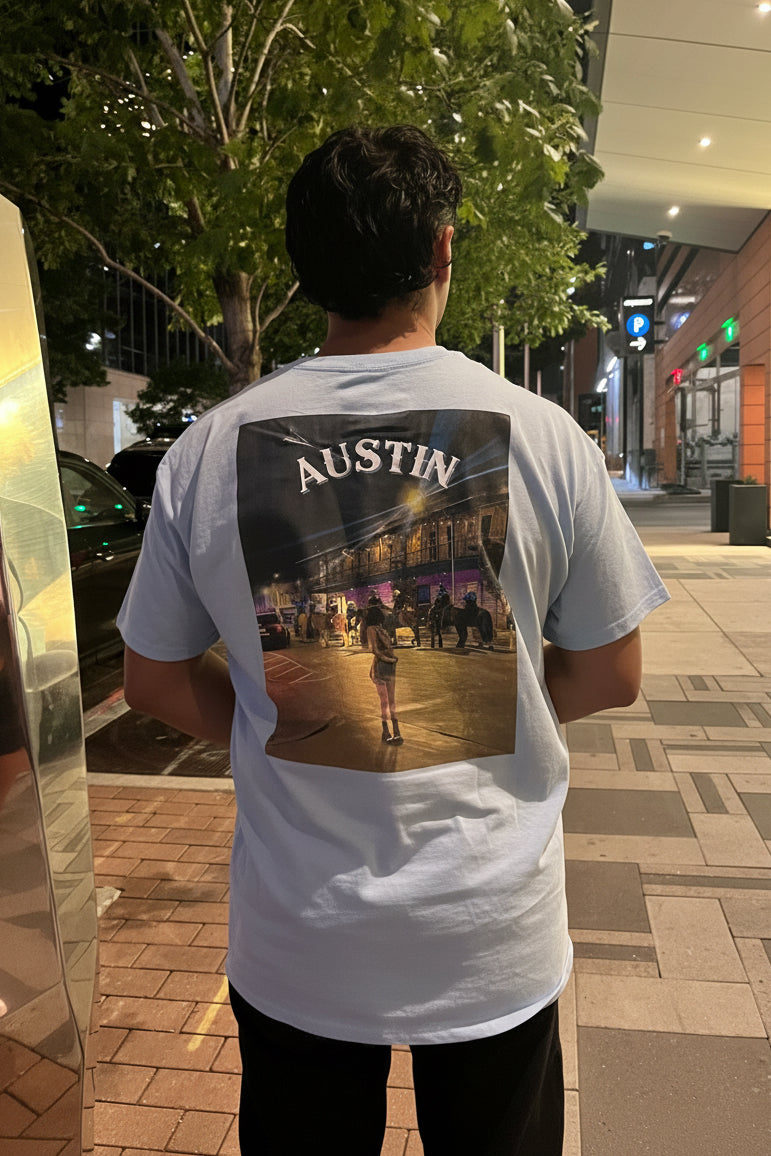 A man viewed from behind wearing a light blue t-shirt with a large graphic print on the back featuring the word 'AUSTIN' and a night scene, standing on a city sidewalk at night.